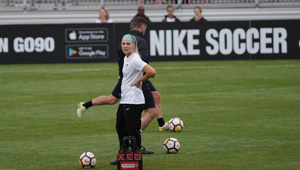 A person in sports attire stands on a soccer field, surrounded by several soccer balls and a carrier with bottles. The field is bordered by advertisements for Nike Soccer and app stores. Several people sit in bleachers in the background.