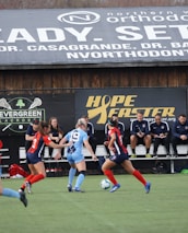 Players engage in a competitive soccer game on a field while others are seated on the bench watching attentively. The action is taking place in front of a wooden structure displaying multiple advertisements, including orthodontics, lacrosse, and field hockey.