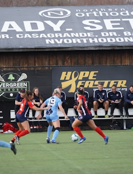 Players engage in a competitive soccer game on a field while others are seated on the bench watching attentively. The action is taking place in front of a wooden structure displaying multiple advertisements, including orthodontics, lacrosse, and field hockey.