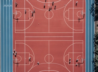 An aerial view of a large, vibrant sports complex featuring a red basketball court with multiple hoops and a surrounding blue running track. Several people are scattered across the area, engaged in various activities.