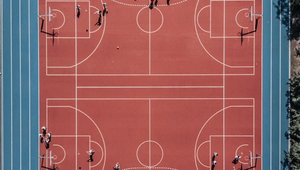 An aerial view of a large, vibrant sports complex featuring a red basketball court with multiple hoops and a surrounding blue running track. Several people are scattered across the area, engaged in various activities.