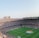 An expansive soccer stadium filled with thousands of spectators. The green field in the center features players and markings, surrounded by vibrant stands and advertising boards on the upper sides of the stadium. The audience is actively watching the game.
