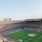 An expansive soccer stadium filled with thousands of spectators. The green field in the center features players and markings, surrounded by vibrant stands and advertising boards on the upper sides of the stadium. The audience is actively watching the game.