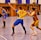 A dynamic indoor sports scene with players in motion on a court. Two athletes in blue and yellow uniforms are engaged in an intense handball game, with one player gripping the other's jersey. The background shows other players, several seated people, and advertisements on the walls.
