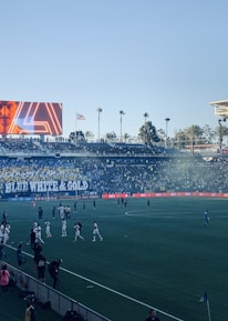 A large sports stadium filled with spectators. The field is occupied by soccer players, some walking and others scattered across the pitch. A massive screen displays a person, and flags fly on top of the stadium roof. The audience is dense, and the atmosphere seems lively with a visible banner showing 'BLUE WHITE & GOLD'.